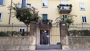 Un edificio amarillo con entrada protegida por una reja, un balcón con plantas en macetas y el número 15 en la reja.