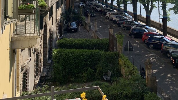 Vista de la calle con coches estacionados, un conjunto de árboles y un edificio con balcones.