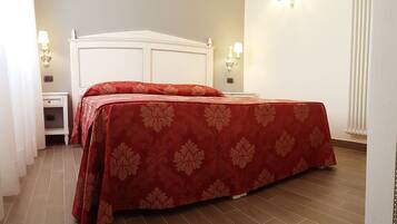 Una cama bien hecha con una colcha roja estampada con motivos florales en una habitación de hotel, con cabecera blanca, dos mesitas de noche y lámparas fijadas a la pared.
