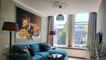 A living room with a teal sofa, a round coffee table, a painting of fruit, a giraffe figurine, and a view of buildings through large windows.