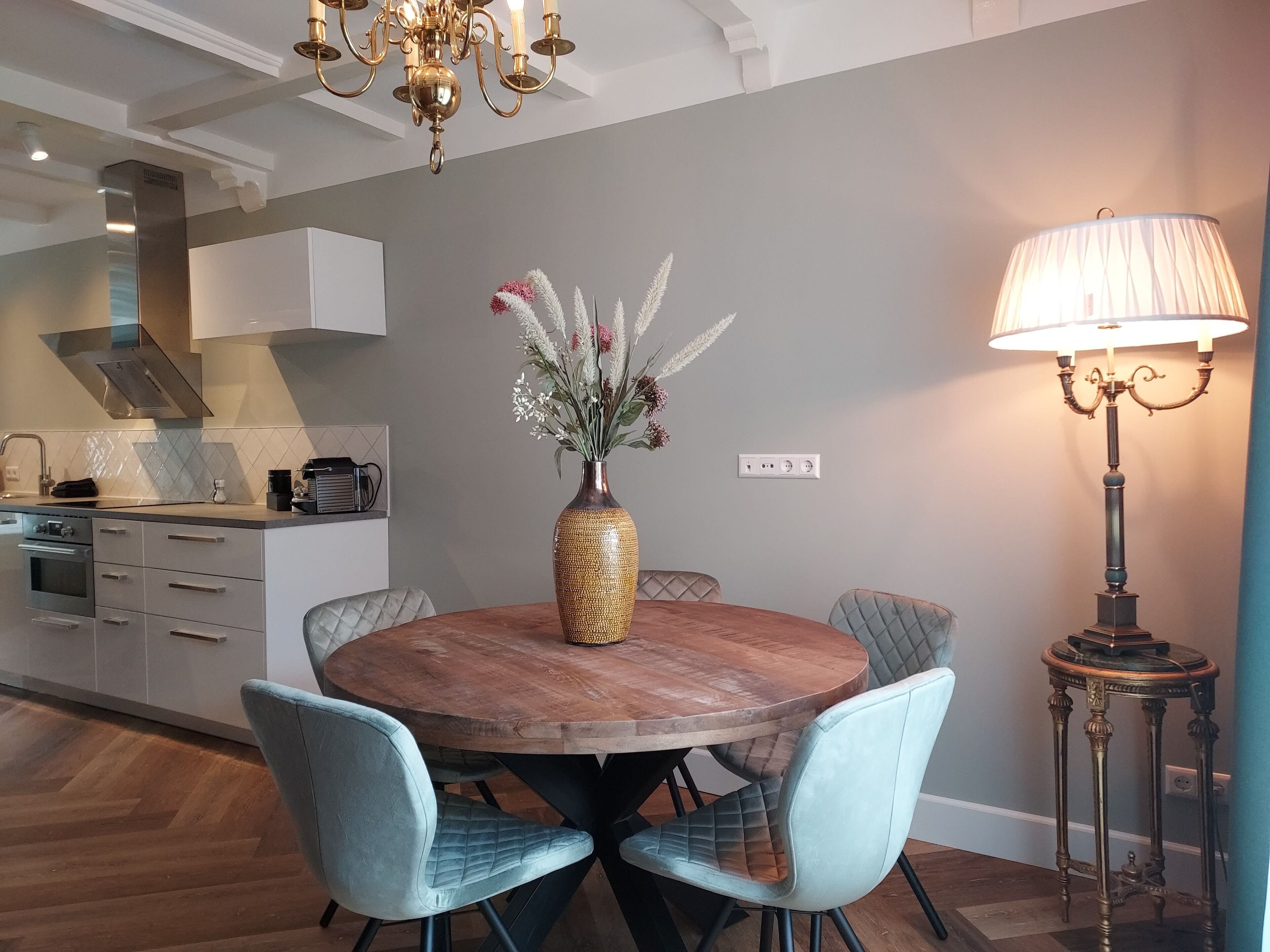 Un comedor con una mesa redonda de madera, dos sillas tapizadas, un florero con flores, una araña y una lámpara de pie.
