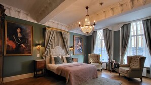 Une chambre spacieuse avec un grand lit, deux fauteuils, un lustre et des œuvres d’art accrochées aux murs.