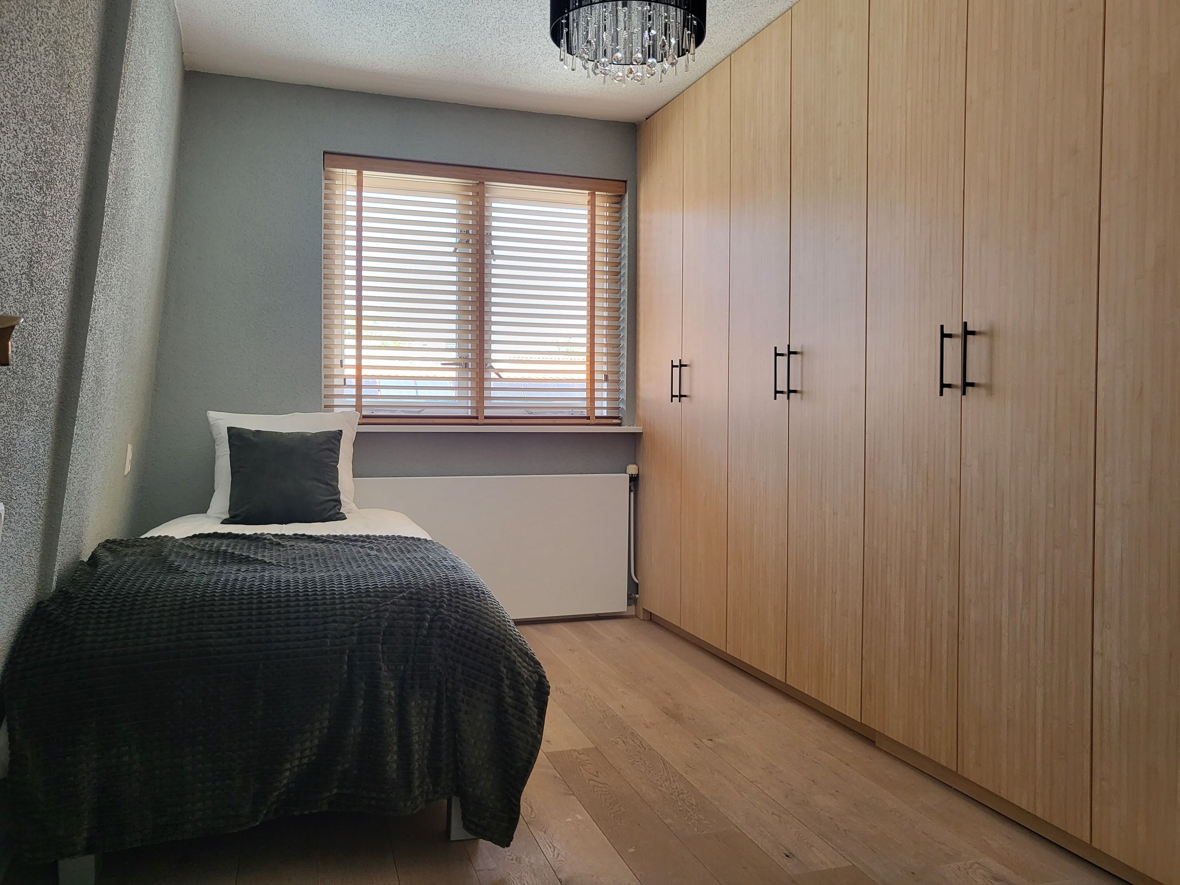 A bedroom with a bed, a window with blinds, a radiator, and a wooden wardrobe.