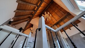 Une pièce aux hauts plafonds, avec des poutres apparentes, un lustre et un escalier moderne doté de balustrades noires.