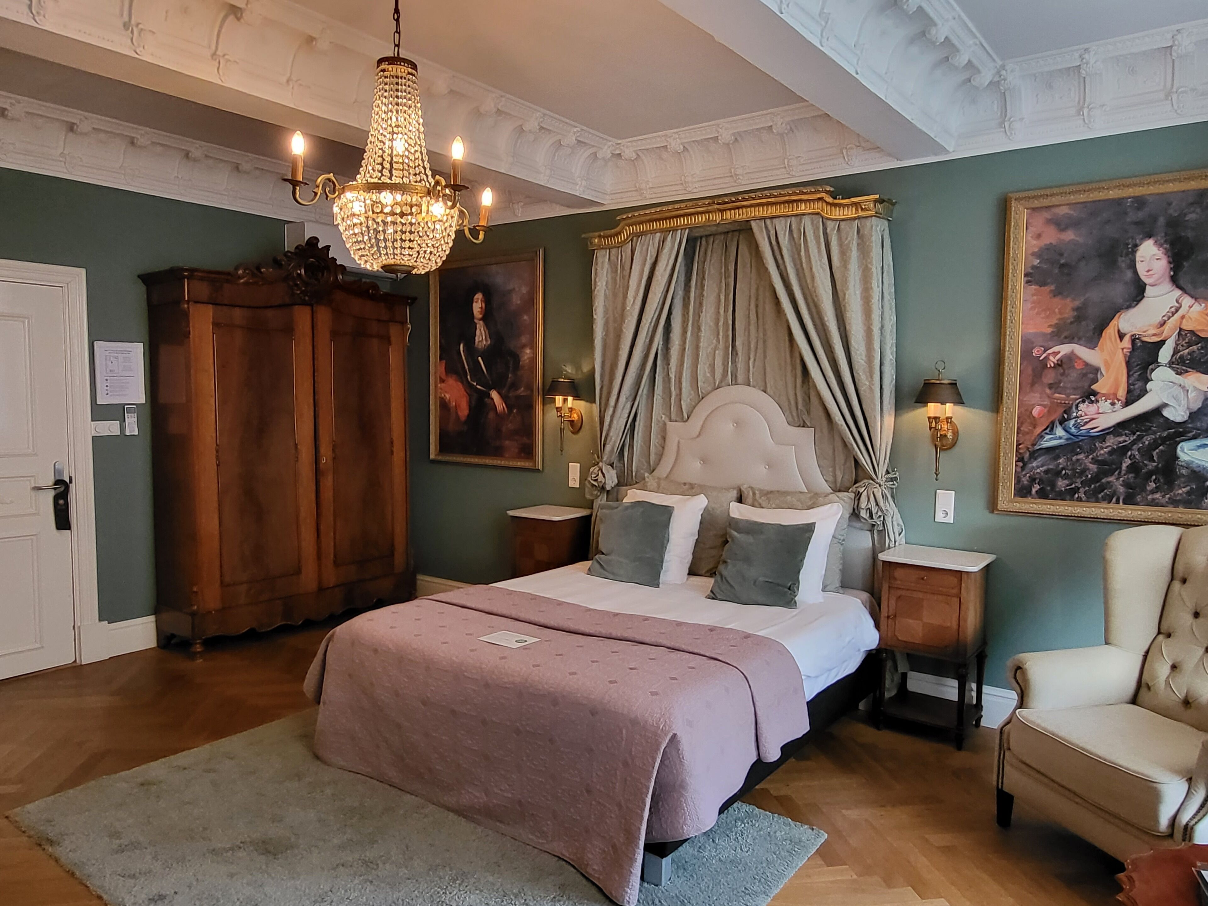Un dormitorio con una cama grande, una araña de cristal, un cuadro, un armario de madera y una silla.
