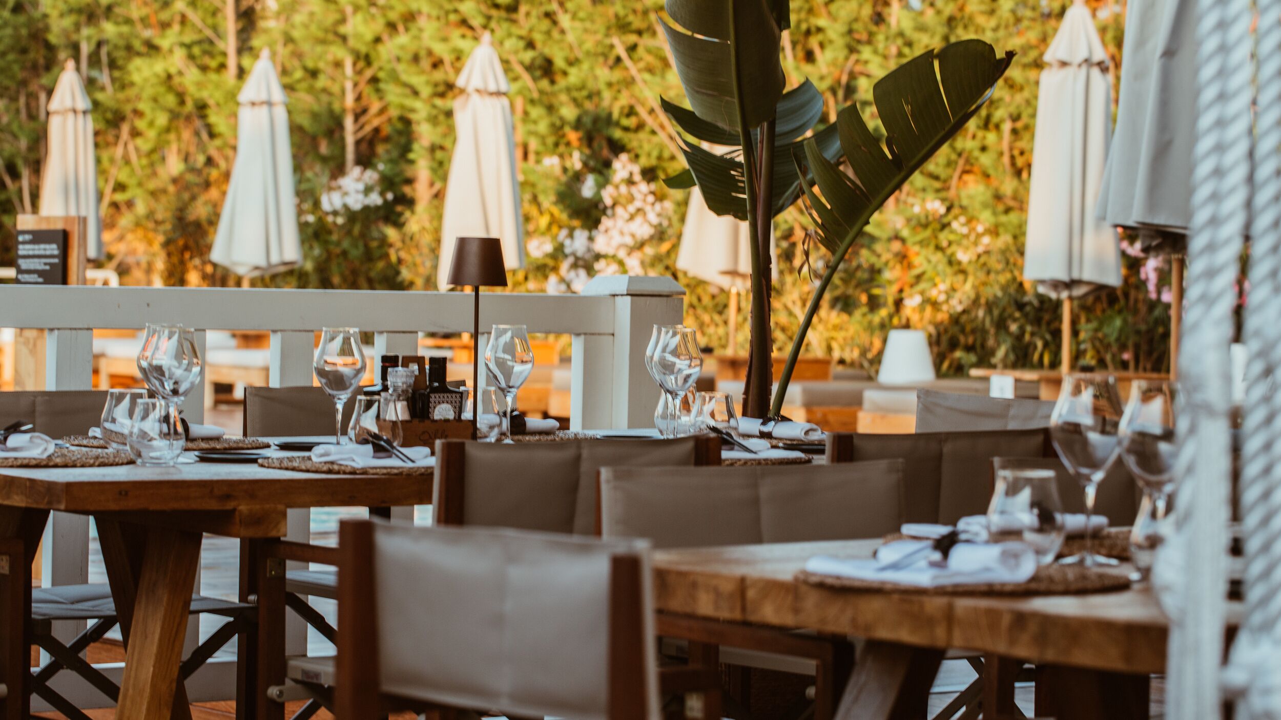 An outdoor dining area with wooden tables and chairs, white umbrellas, and a backdrop of greenery.