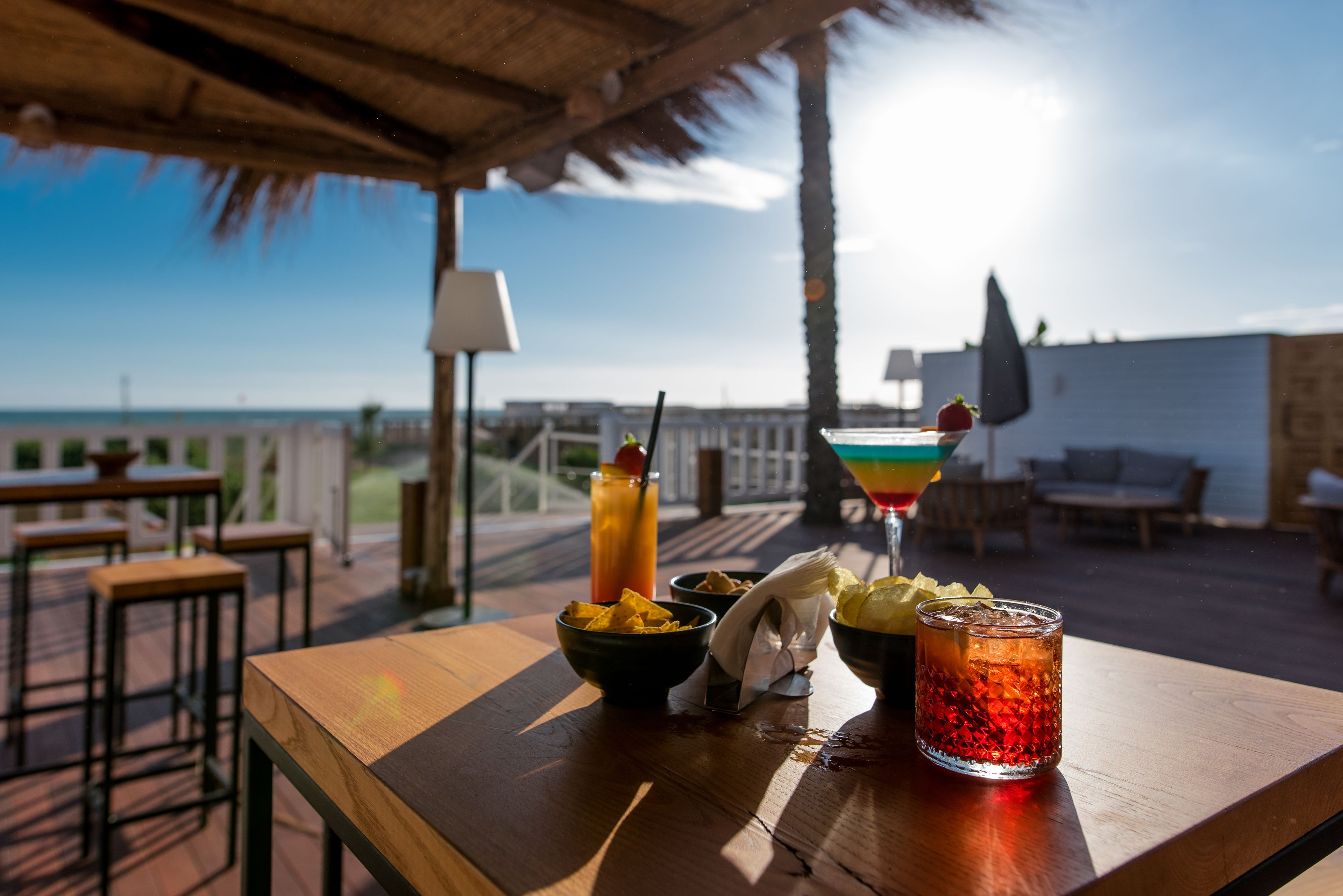 A wooden table with a drink, a bowl of chips, and a napkin on a deck with a view of the ocean.