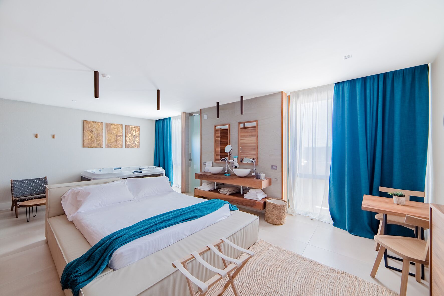 A modern hotel room with a large bed, a sitting area with a chair and table, a bathroom with a vanity and mirror, and a view of the outdoors through large windows.