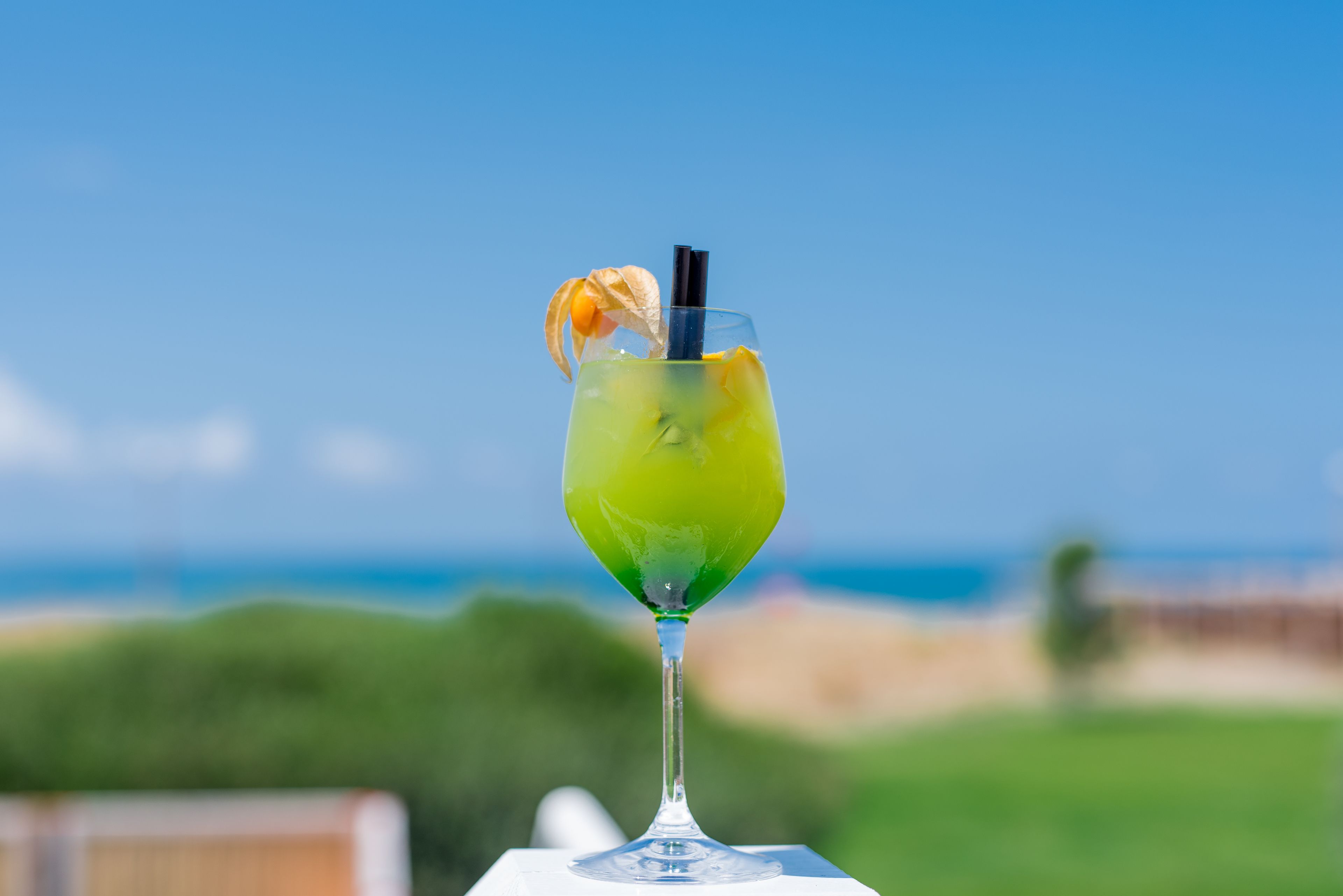 Ett cocktailglas med en grön drink, ett svart sugrör och en orangefärgad dekoration, placerat på ett vitt bord med en suddig utomhusmiljö i bakgrunden.