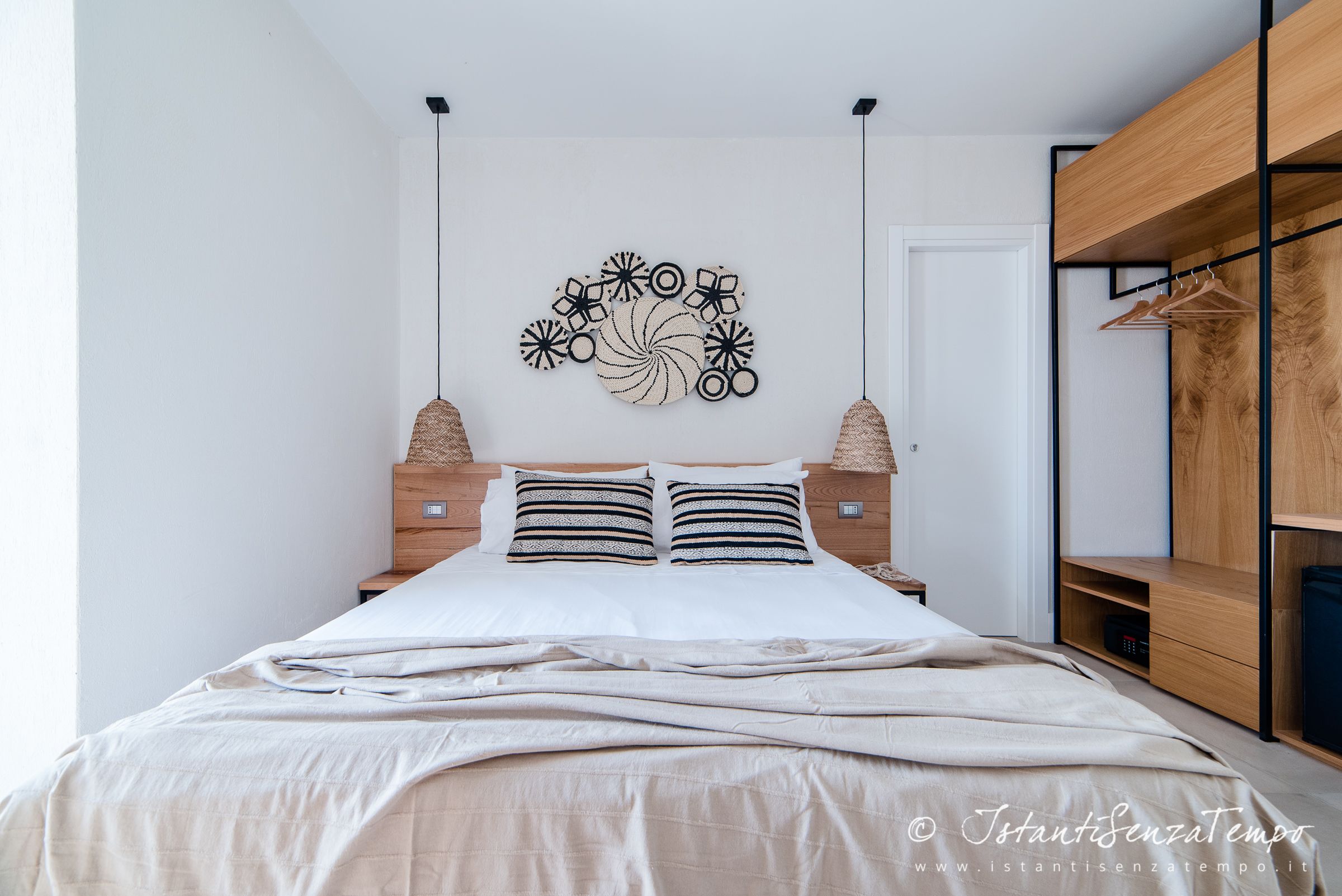 A neatly made bed with a beige comforter, a wall-mounted abstract art piece, and a wooden wardrobe with hanging space.