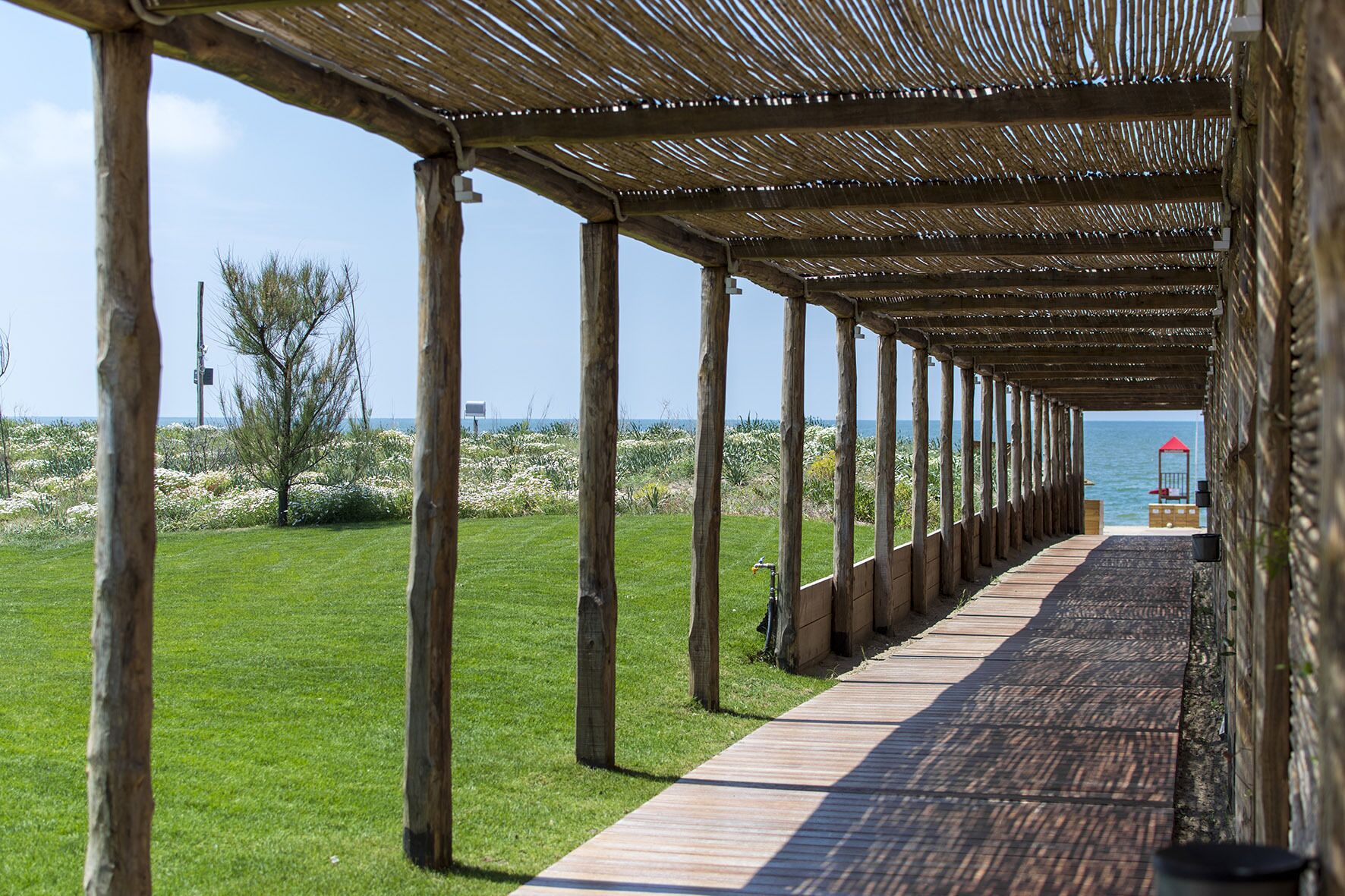 A wooden deck leading to a beach with a covered area and a lifeguard tower in the distance.