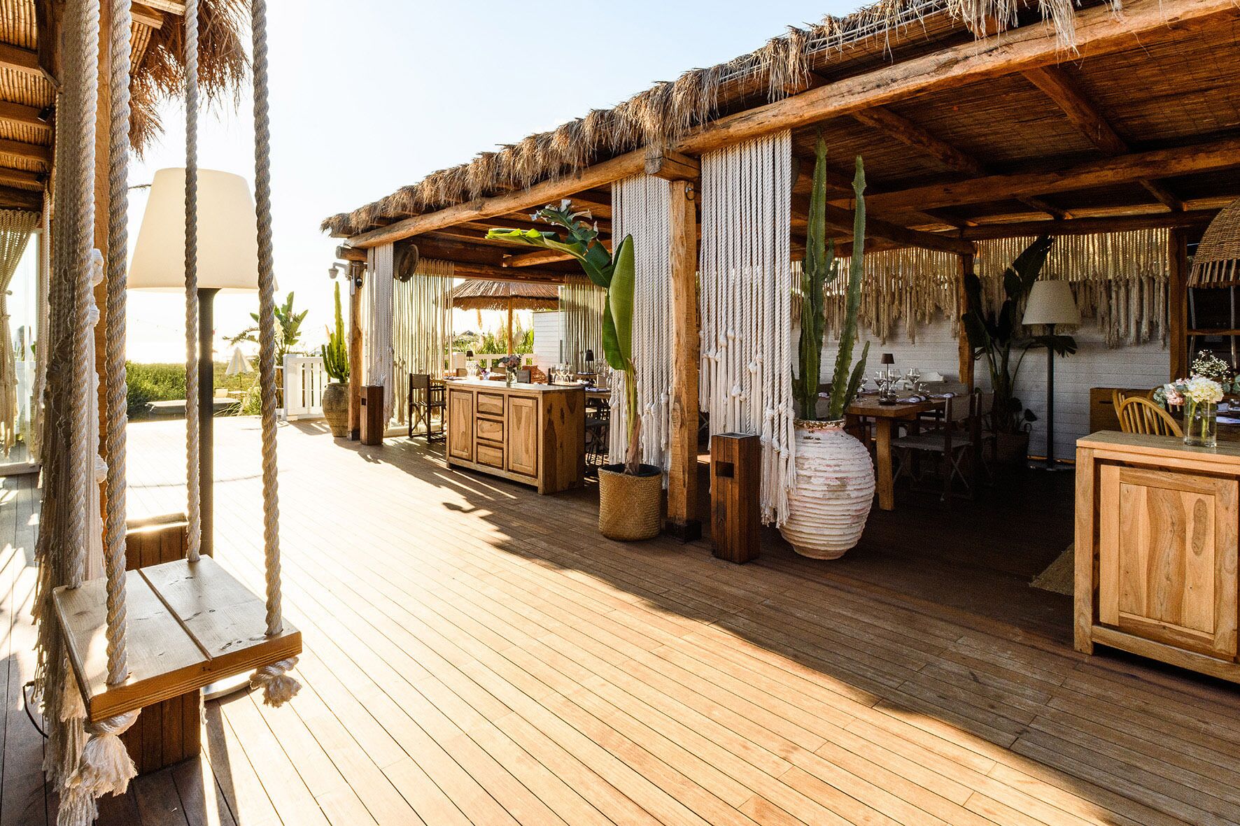 A wooden deck with a thatched roof, wooden furniture, and hanging plants.