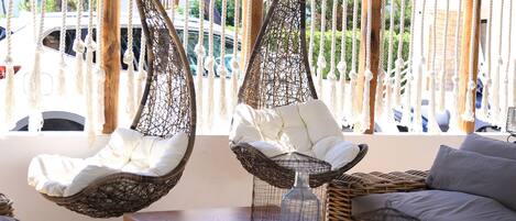 Dos sillones colgantes con cojines blancos, un sofá de mimbre y una pequeña mesa con un florero de cristal.