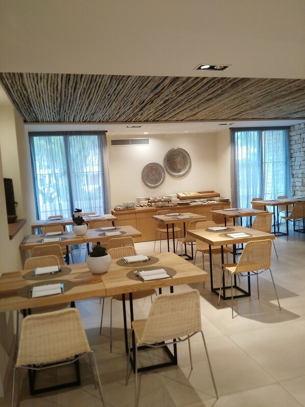 A modern dining area with wooden tables and chairs, large windows with sheer curtains, and decorative plates on the wall.