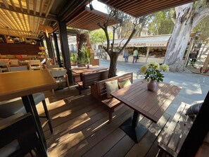 An outdoor seating area with wooden tables and benches, surrounded by trees and a covered walkway.