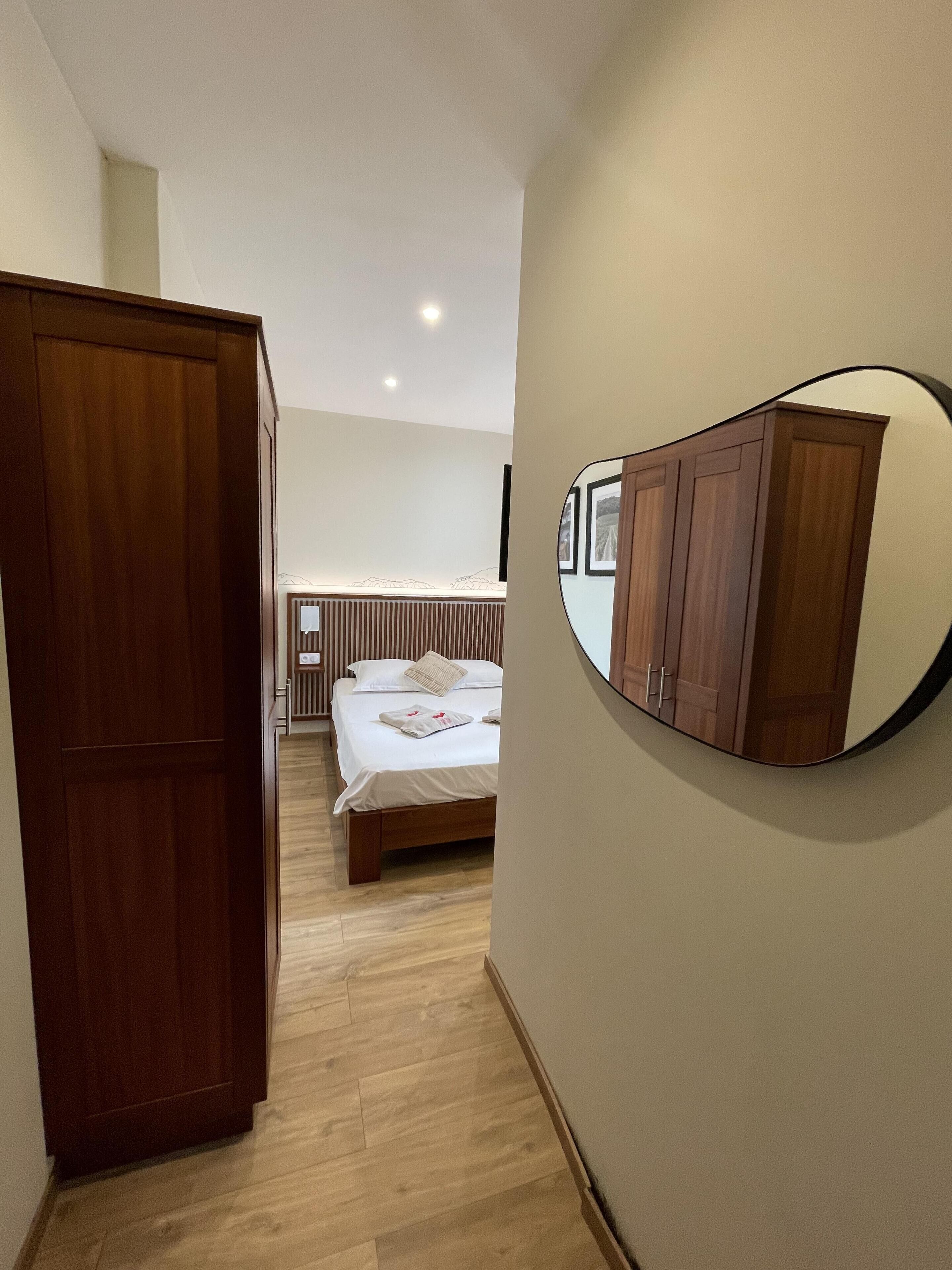 Ein Schlafzimmer mit einem hölzernen Kleiderschrank, einem Bett mit weißer Bettwäsche, einem Nachttisch und einem an der Wand befestigten Spiegel, der den Raum reflektiert.