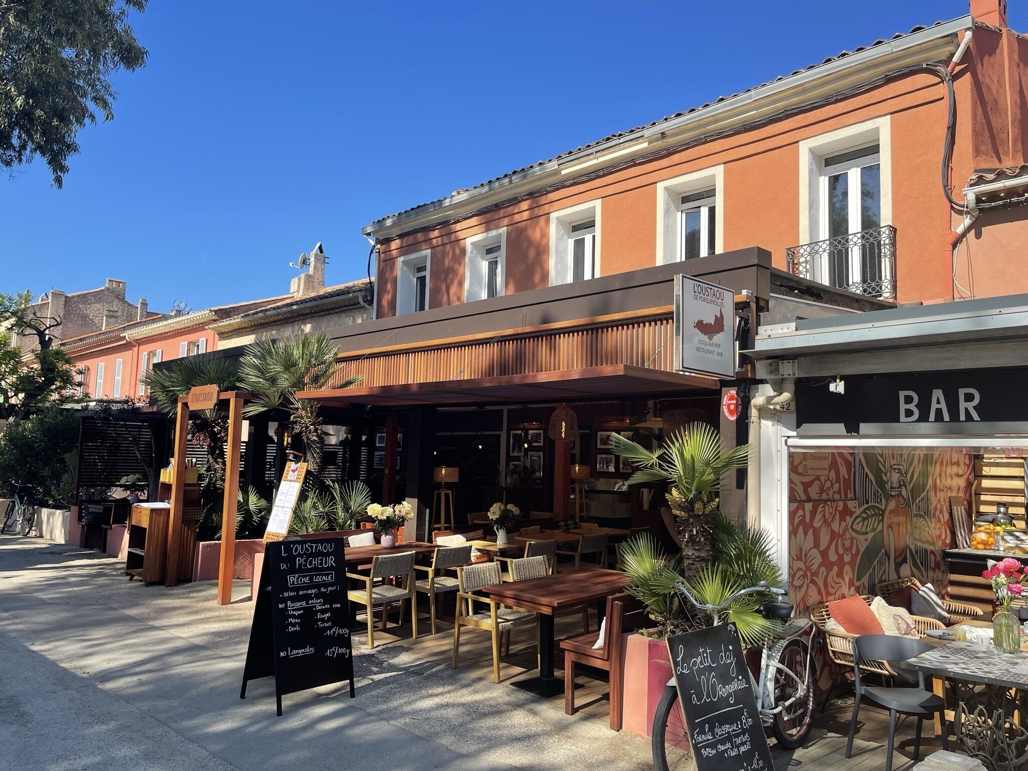 An outdoor seating area with wooden tables and chairs, a sign for 'L'OUSTAOU DU PECH', and a 'BAR' sign.