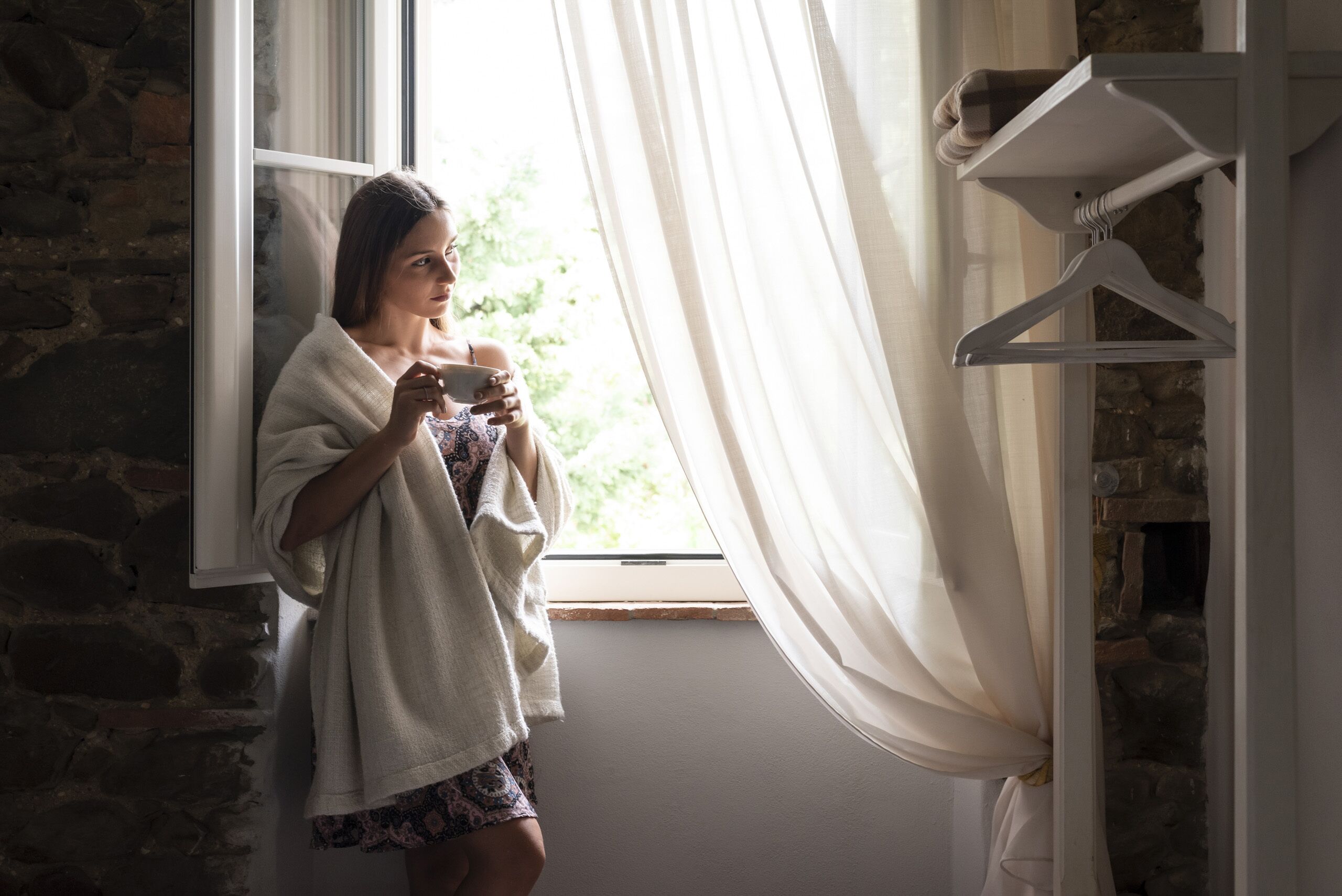 A person standing by a window with sheer curtains, holding a camera.