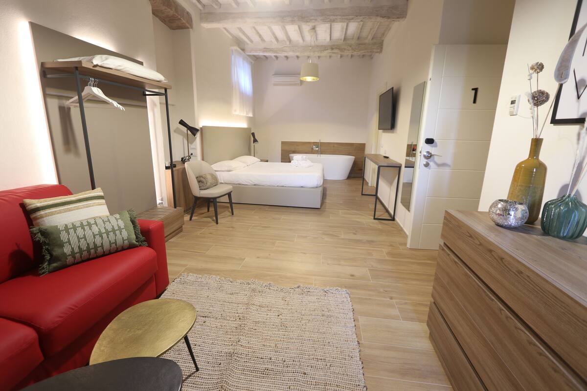 A modern hotel room with a red sofa, a large bed, a wooden dresser, and a bathroom with a bathtub.