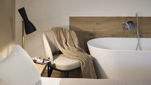 A modern bathroom with a freestanding bathtub, a chair with a beige throw blanket, and a floor lamp. - Botrona B&B (Scarlino)