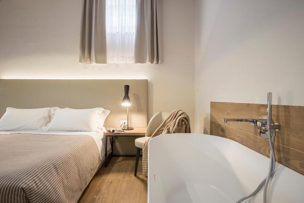 A hotel room with a bed, bedside table, lamp, chair, and a bathtub.