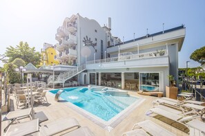 Un hotel con una amplia piscina exterior, varias zonas de descanso y un edificio distintivo con balcones y el logotipo de una corona.