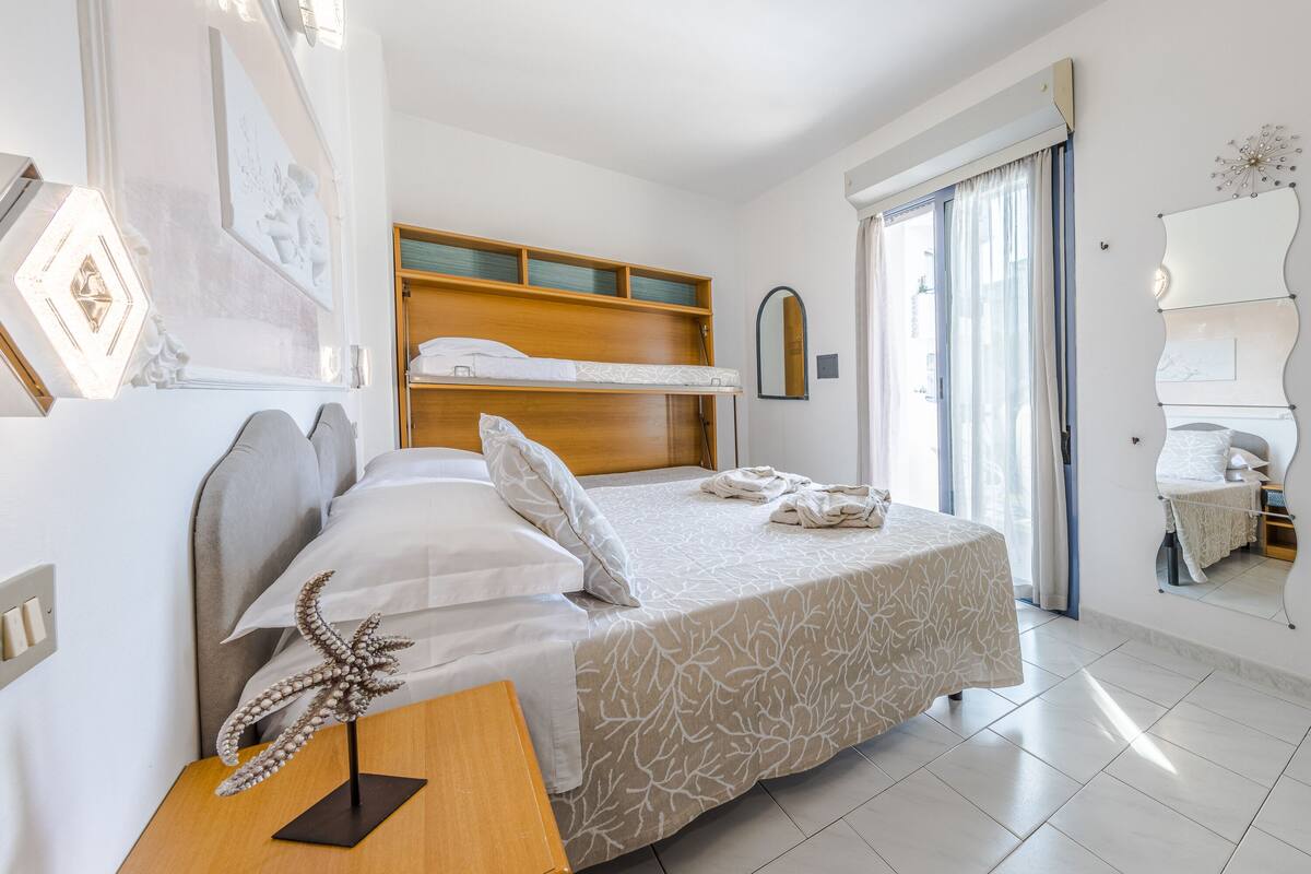 Habitación de hotel con literas, una mesita de noche con lámpara, un espejo grande y una ventana con cortinas.