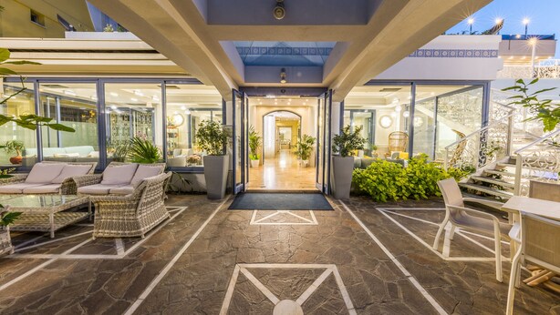 Un hall d’hôtel moderne avec un sol en pierre, des meubles en osier et une entrée en verre donnant sur un escalier.