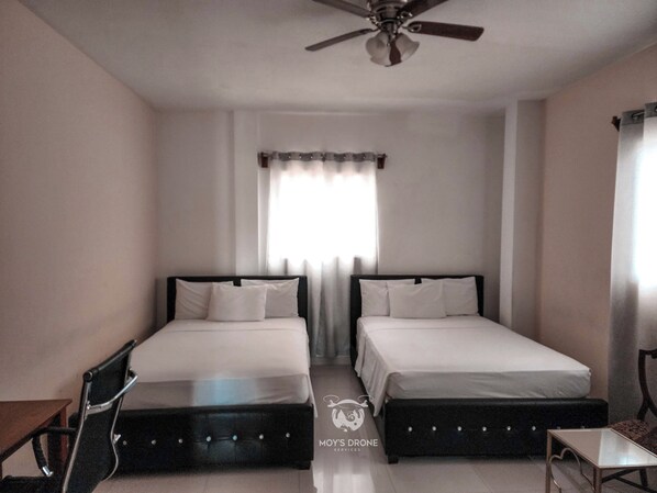 A hotel room with two beds, a ceiling fan, a window with curtains, and a small table.