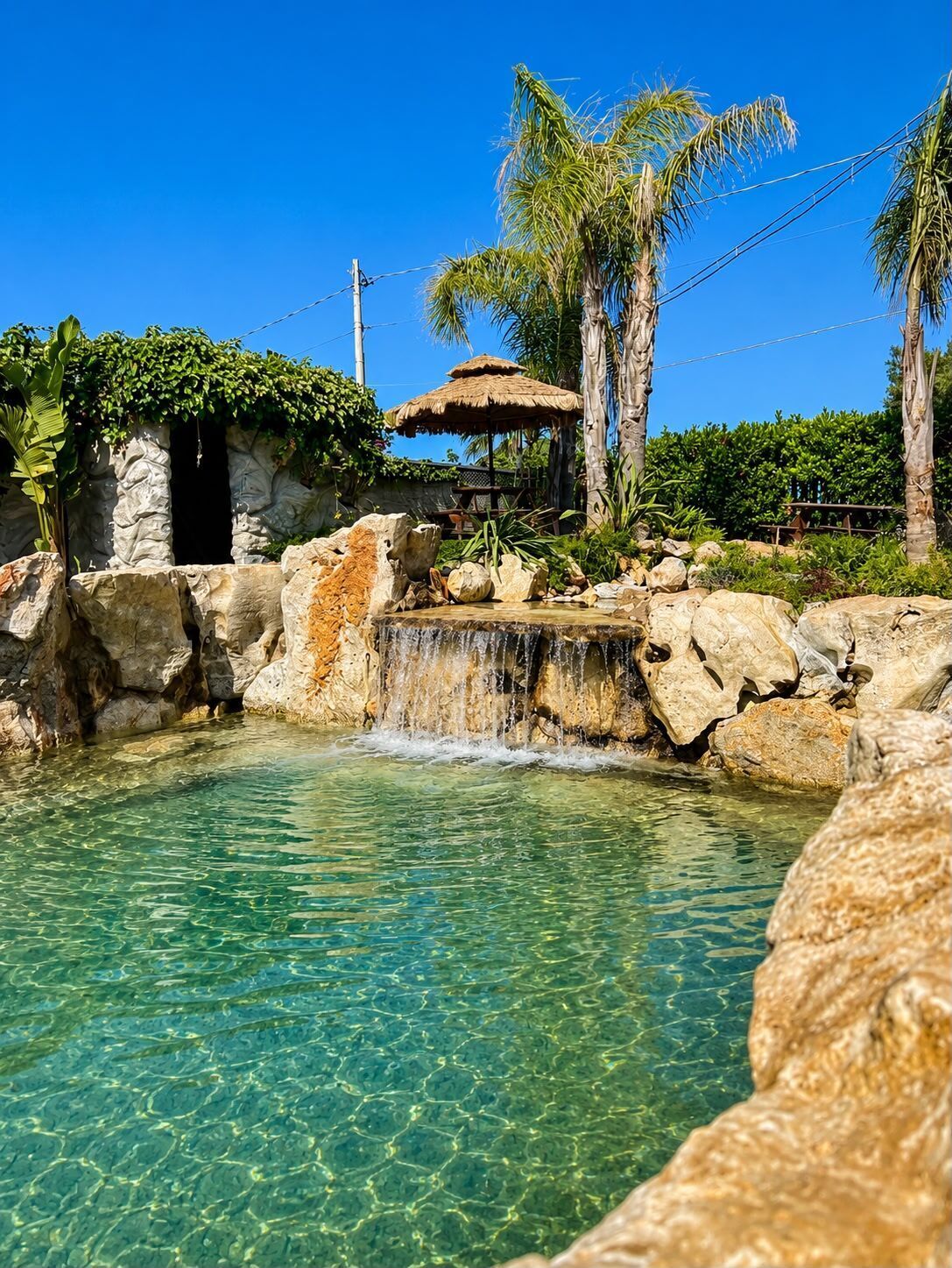 A clear pool with a waterfall, palm trees, and a rustic hut.
