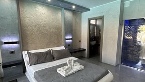 A modern hotel room with a large bed, a bathroom with a glass door, and a stone wall feature. - Villa Rosa dei Venti (Cinisi)
