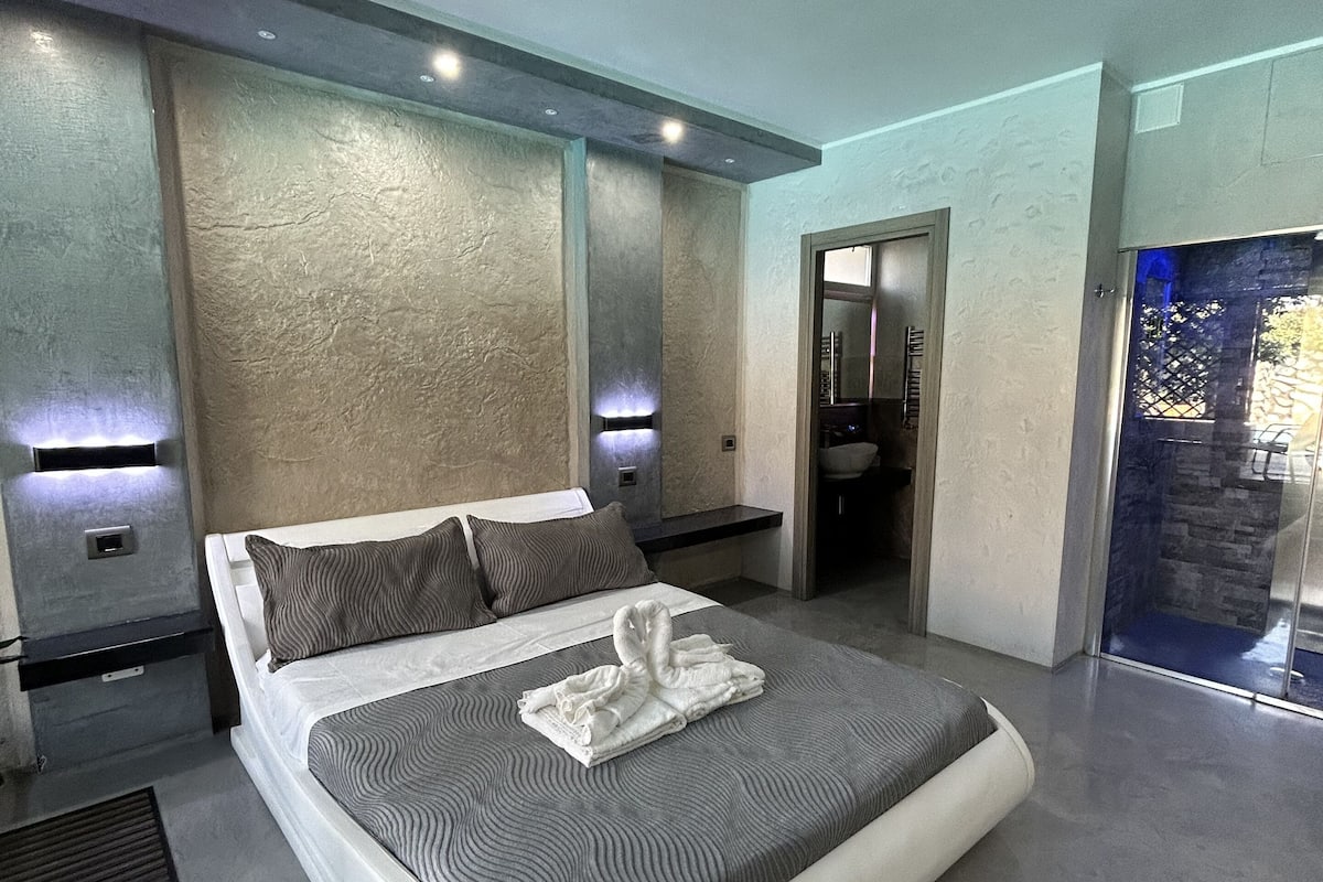Ein modernes Hotelzimmer mit einem großen Bett, einem Badezimmer mit Glasdusche und einer Wandverkleidung aus Stein.