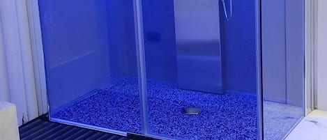 A glass-enclosed shower with a blue light, a metal door, and a drain.