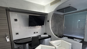 A modern hotel room with a curved glass partition, a flat-screen TV, a small desk, two white leather sofas, and a small round table with a plant. - Villa Rosa dei Venti (Cinisi)