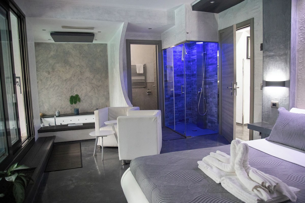 Ein modernes Hotelzimmer mit einem großen Bett, einer gläsernen Dusche, einer Sitzecke mit einem Sessel und einem kleinen Tisch.