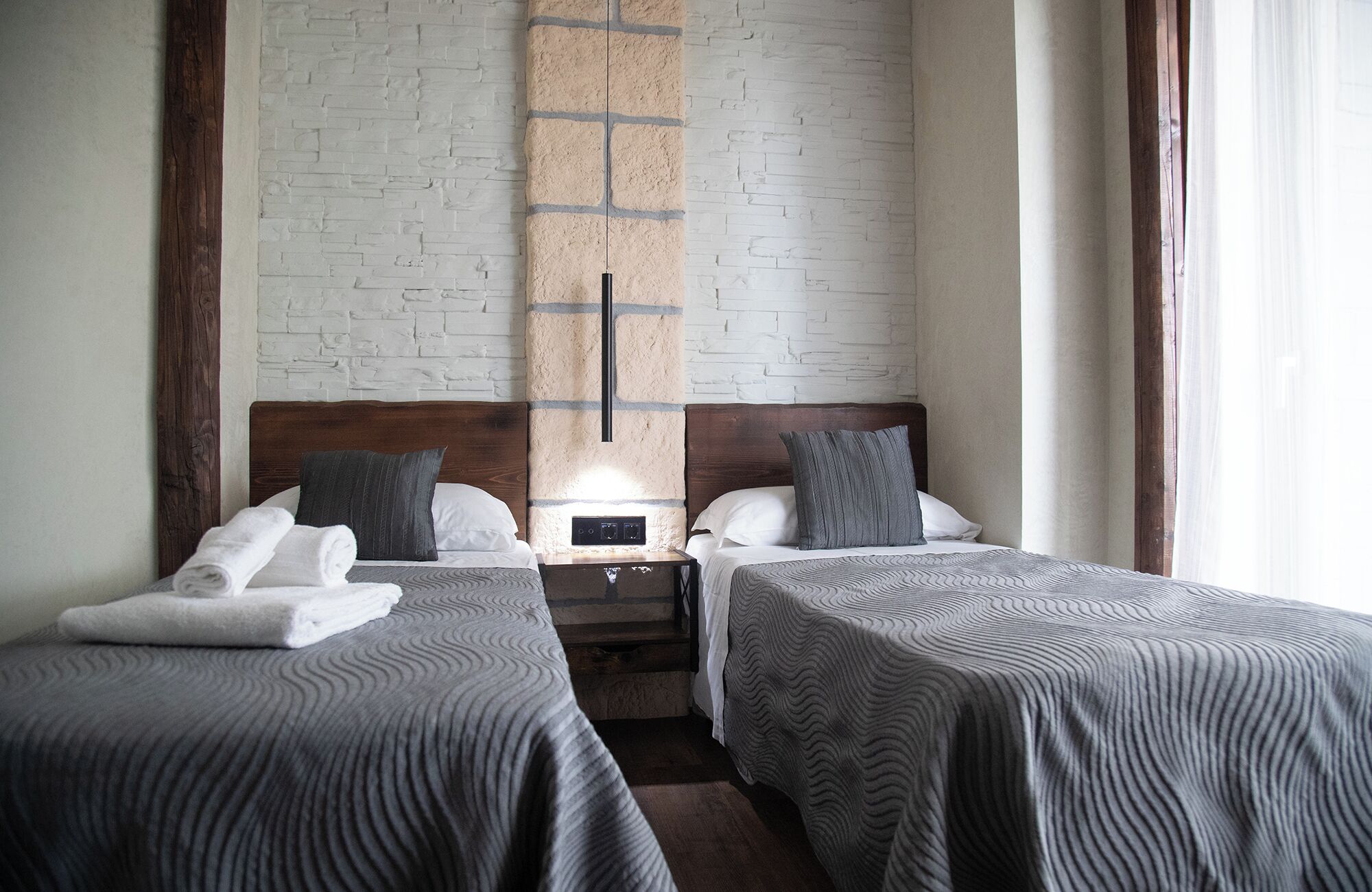 A hotel room with two single beds, a brick-patterned wall, a wooden headboard, and a nightstand with a lamp.