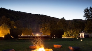 A campfire with logs and rocks in a grassy field, surrounded by trees and a building with lights in the background. - The Graham & Co. (Phoenicia)