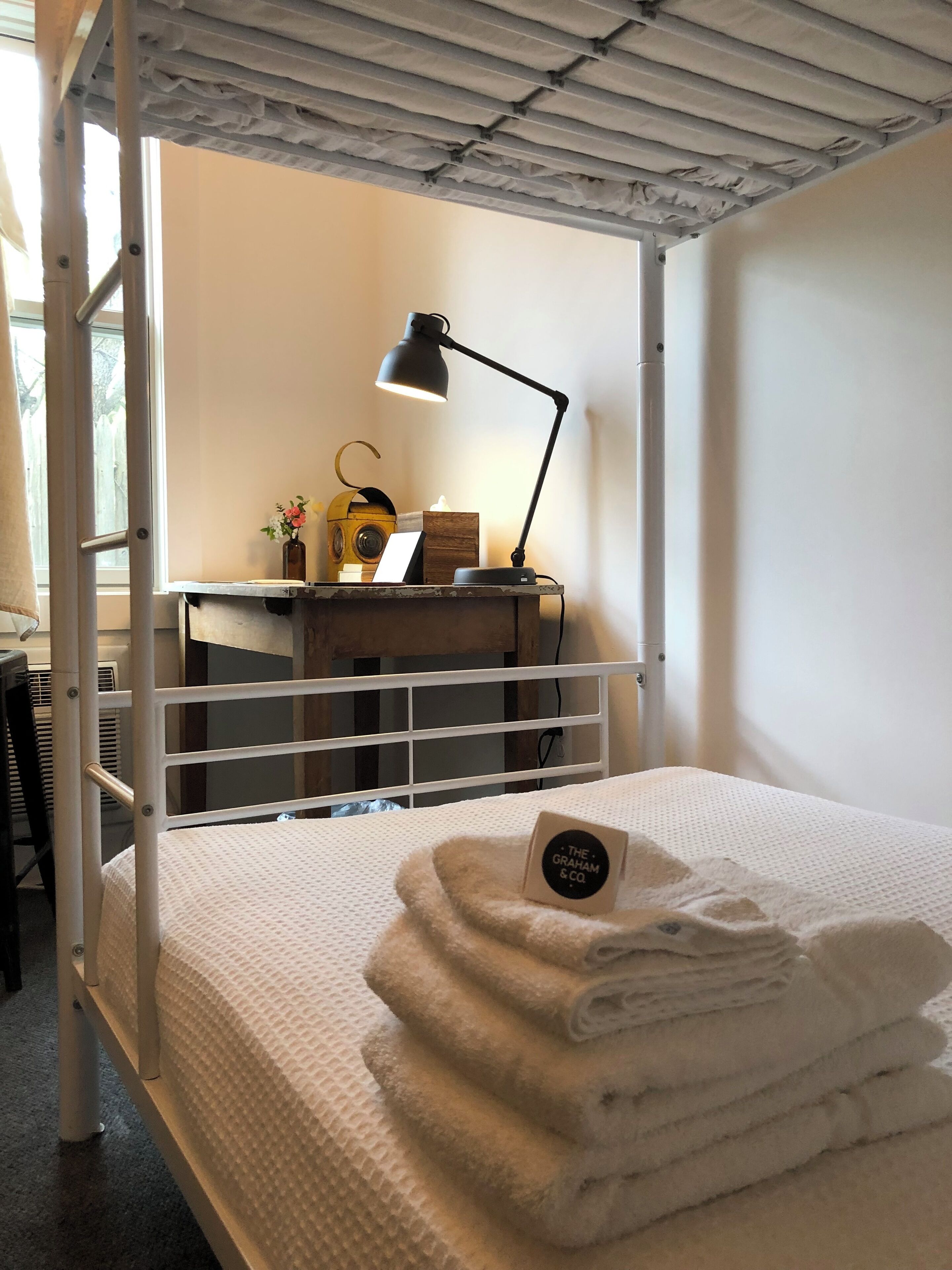 Een stapelbed met een wit matras en een klein bureau met een lamp. Er hangt een bordje aan het bed en er is een raam zichtbaar op de achtergrond.
