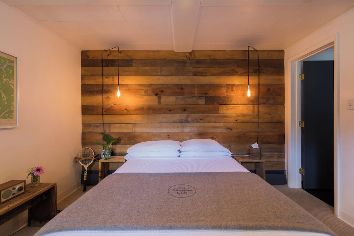 A bedroom with a wooden headboard, a bed with a branded cover, a bedside table with a radio, and a door leading to another room.