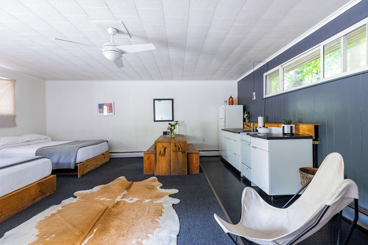 A modern bedroom with two beds, a wooden coffee table, a white chair, and a kitchenette.