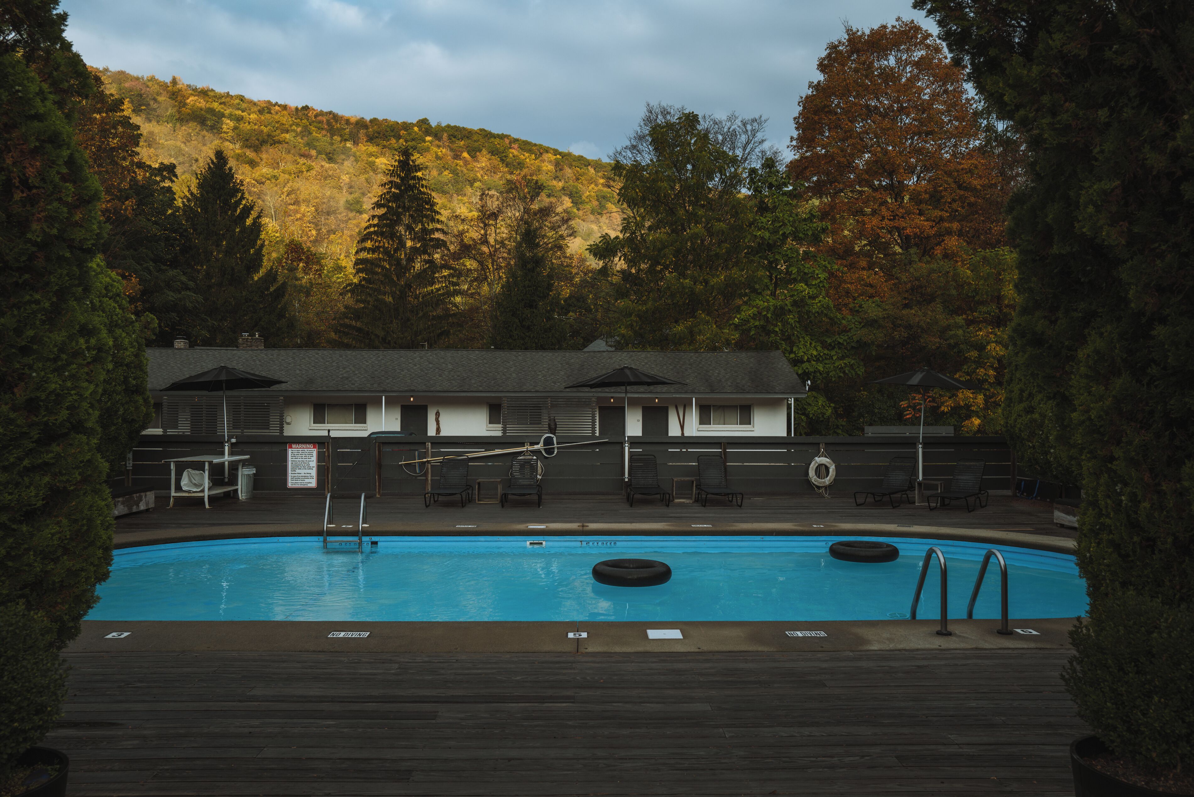 Piscina com espreguiçadeiras e uma boia, cercada por árvores e um prédio com telhado inclinado.