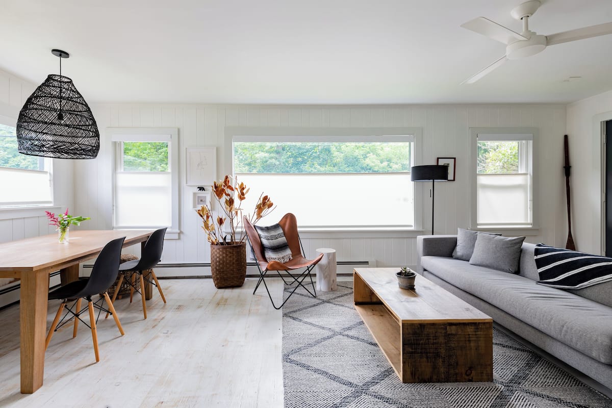 A modern living room with a wooden dining table, black chairs, a grey sofa, and a ceiling fan.