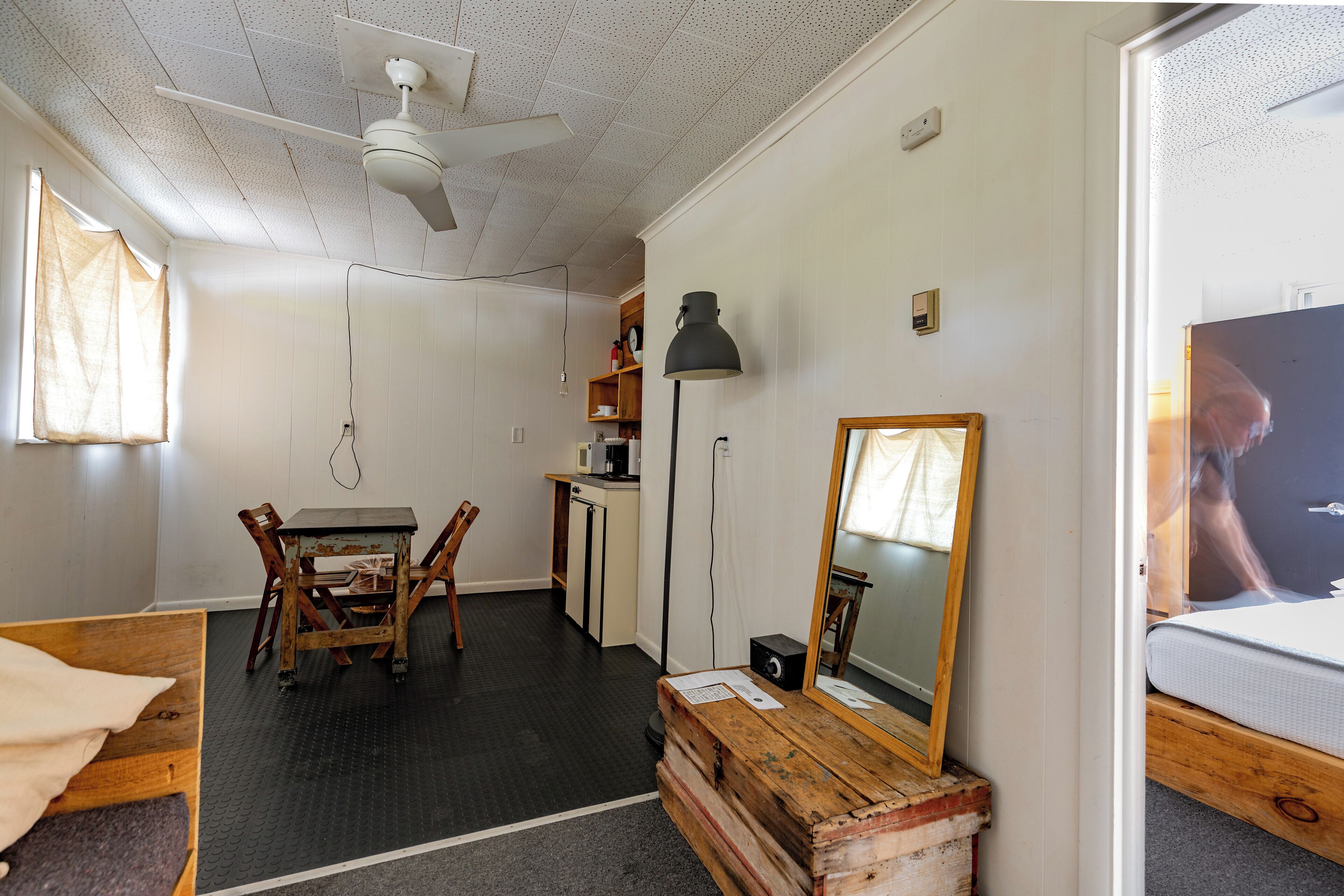 Een kleine kamer met een bed, een houten bureau, een stoel, een kitchenette en een plafondventilator.