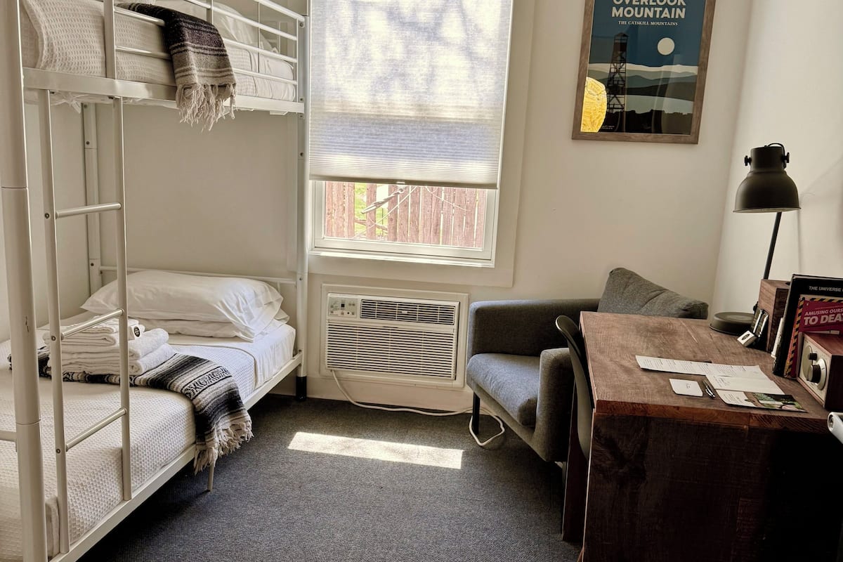 A dormitory room with bunk beds, a desk, a chair, a radiator, and a window with a view of a fence.