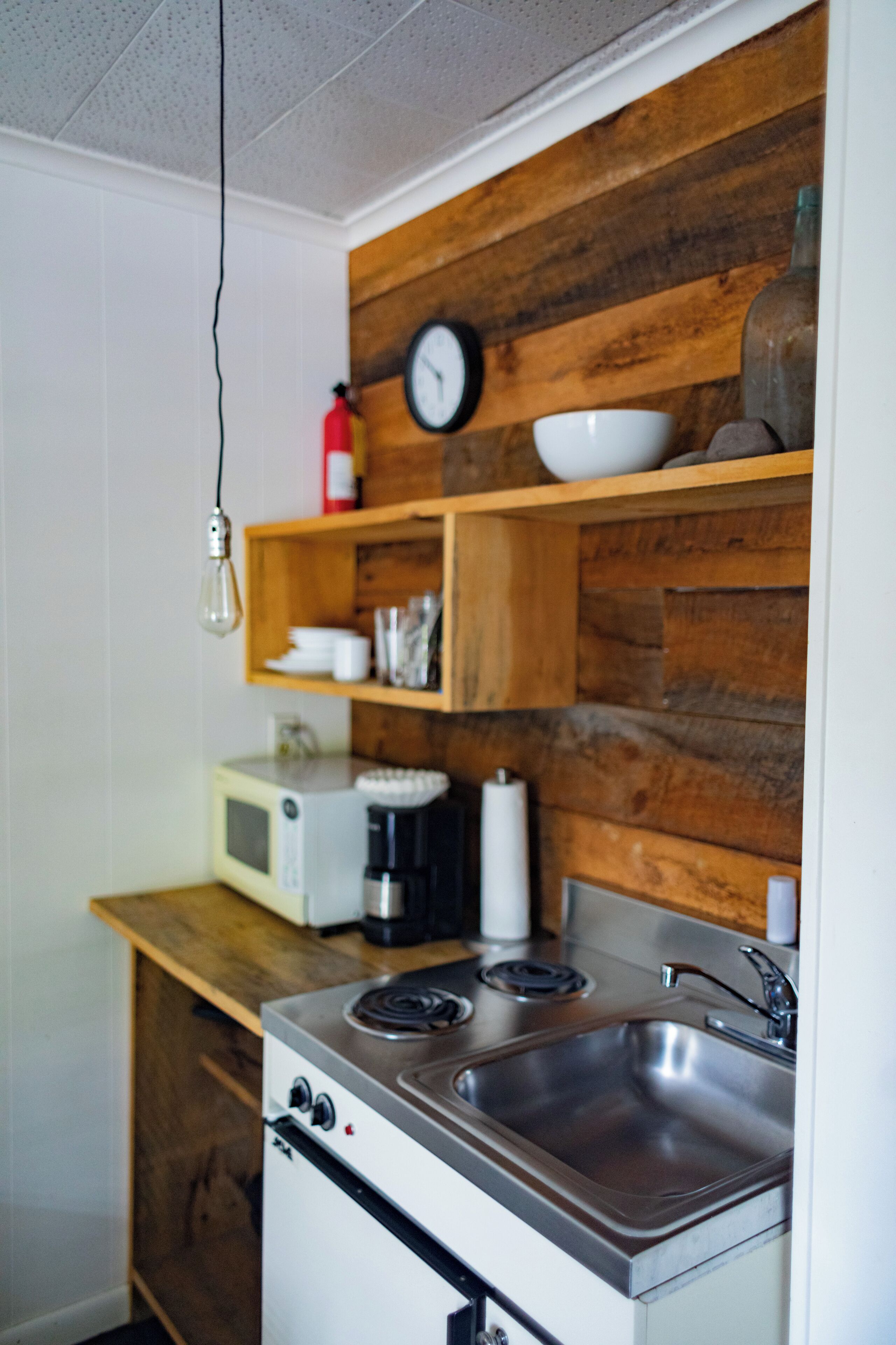 Cocina pequeña con microondas, estufa y lavamanos. Hay un estante de madera sobre la estufa con un reloj y un bol. Una bombilla cuelga del techo.