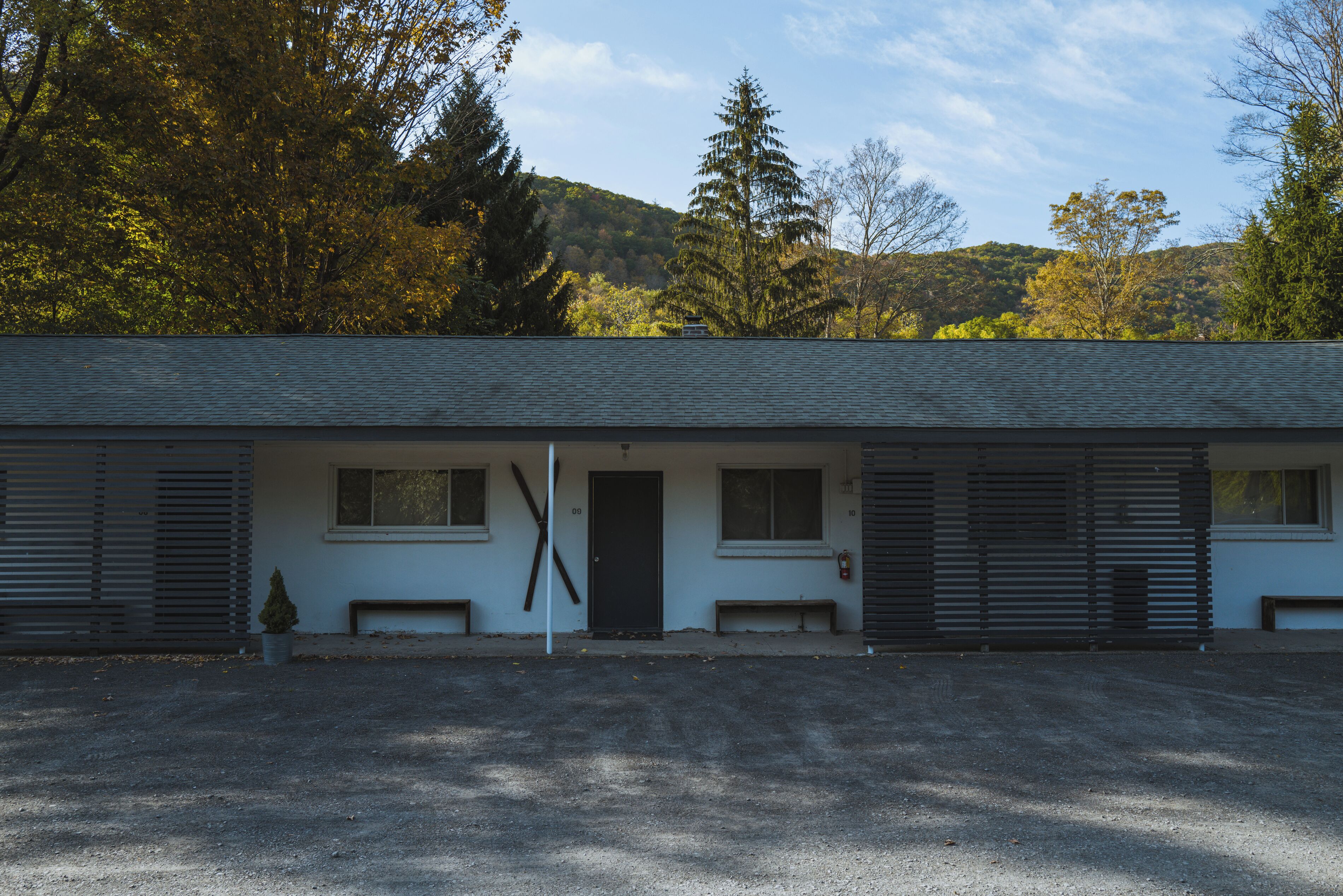 A building with a sloped roof, multiple windows, and a door. There are benches outside and a small tree in front.