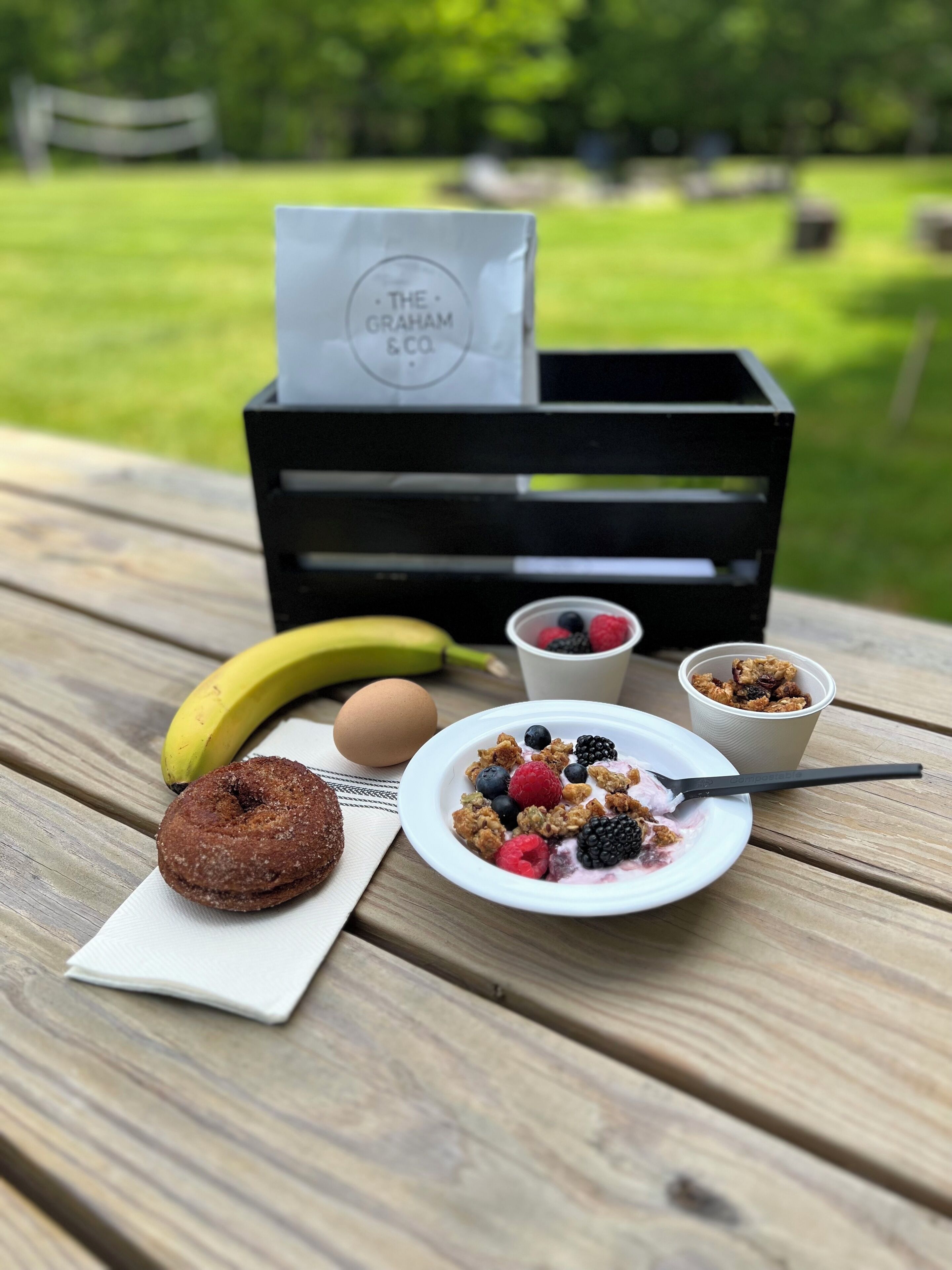 Une table en bois dressée pour un pique-nique, avec une assiette de fruits rouges et de céréales, une banane, un œuf et un beignet.