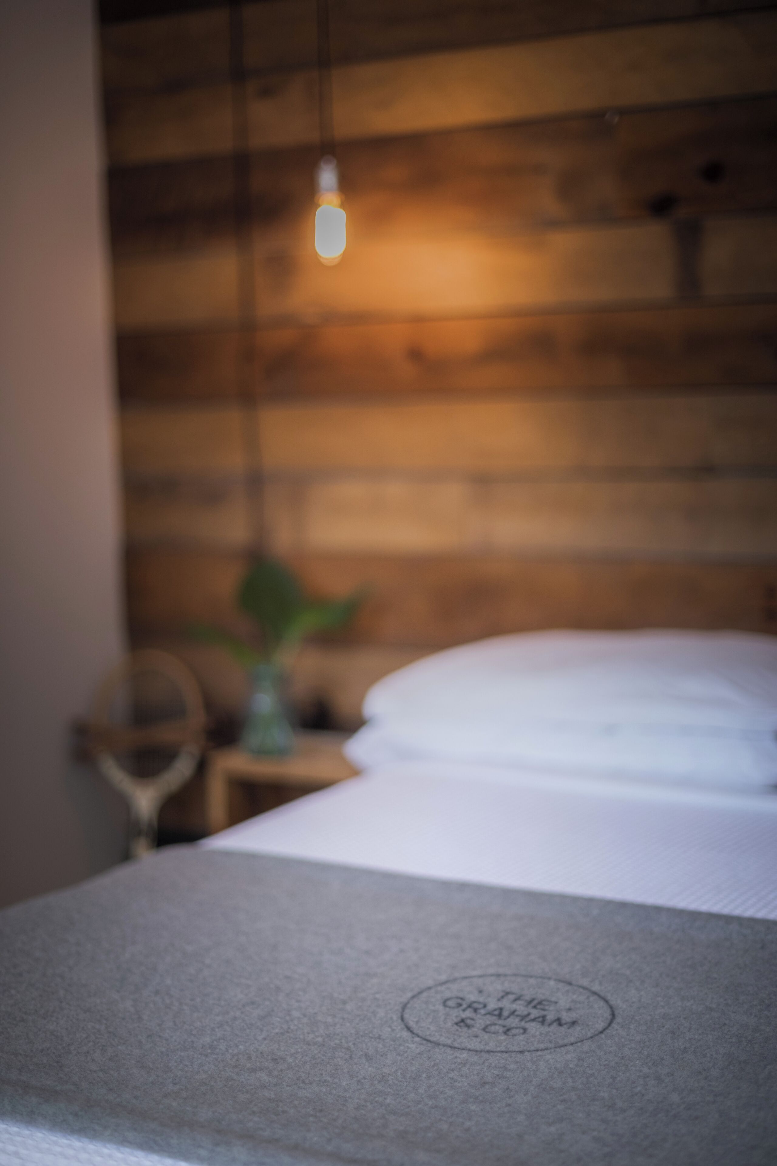 Un lit avec un drap blanc et un logo sur la tête de lit. Un luminaire suspendu se trouve au-dessus du lit.