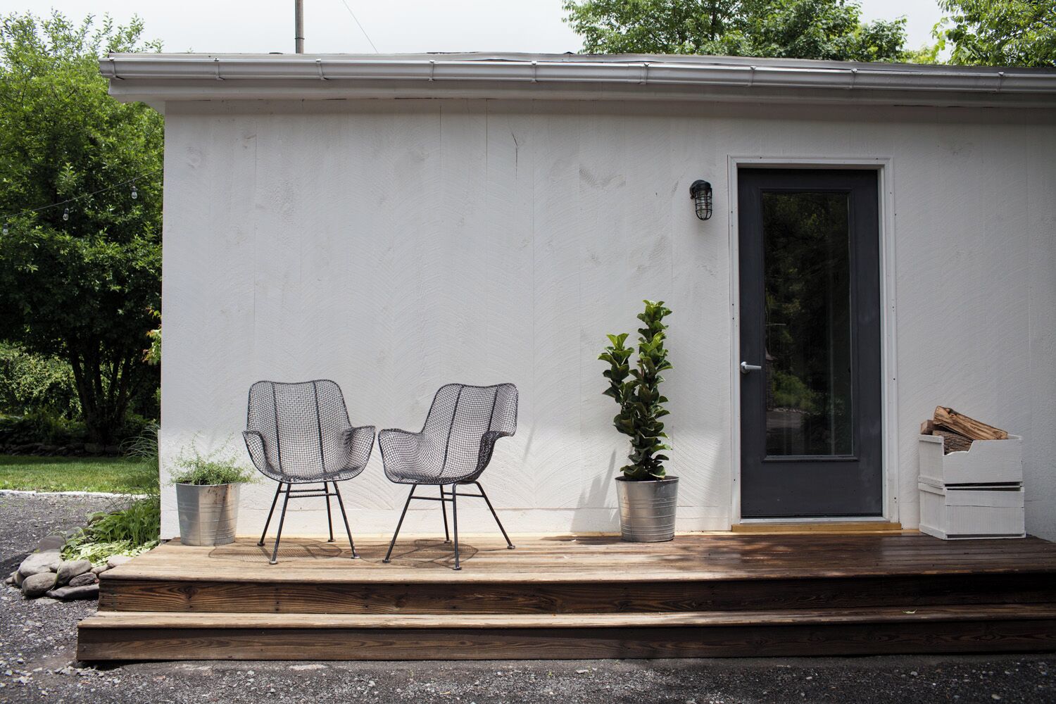A small, single-story building with a white exterior, a dark door, and a small porch area with two wireframe chairs and a potted plant.