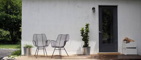 A small, single-story building with a white exterior, a dark door, and a small porch area with two wireframe chairs and a potted plant.